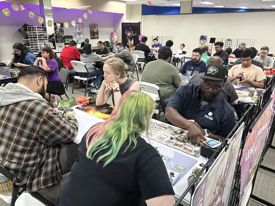 A packed event night in Game Nook's play space, with roughly twenty-five players at tables running trading card game matches.