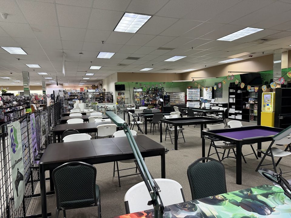 Game Nook's play space set with Box King gaming tables and chairs, one table topped with a purple play surface, magnifier lamps in the foreground and the shop's shelves beyond.
