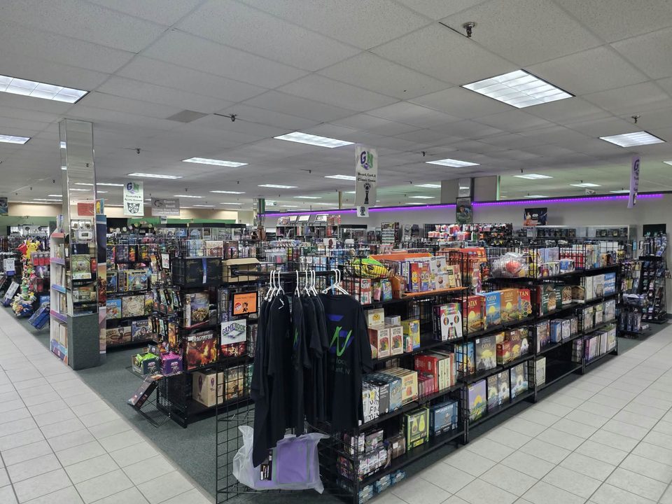 Game Nook's retail floor: board games including Catan and Exploding Kittens on wire shelving, Game Nook T-shirts on a rack, and department signs hanging overhead.