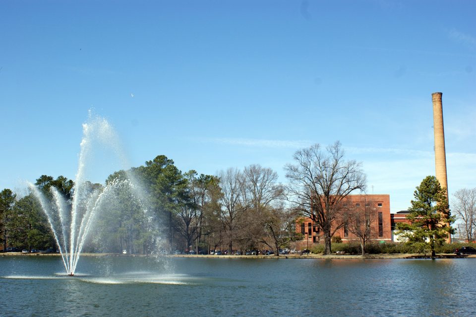 Rocky Mount City Lake on a clear day, with a fountain spraying in the foreground, trees along the shore and a brick building and tall smokestack beyond.